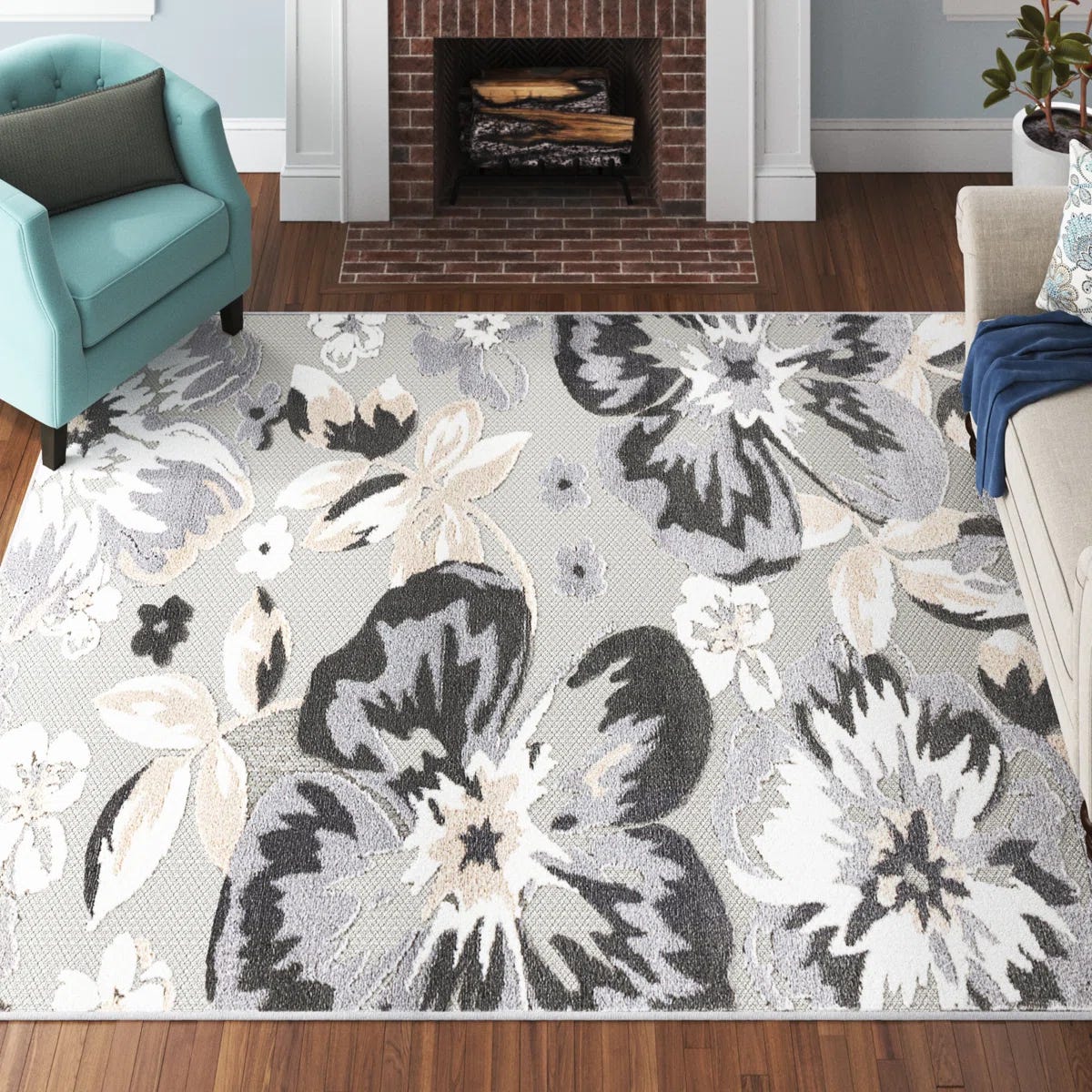 A living room features a large area rug with a floral pattern in shades of gray, white, and beige, set on wooden flooring. An aqua armchair and light-colored sofa flank a brick fireplace.