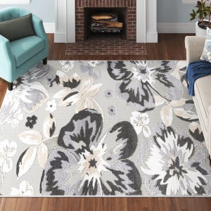 A living room features a large area rug with a floral pattern in shades of gray, white, and beige, set on wooden flooring. An aqua armchair and light-colored sofa flank a brick fireplace.