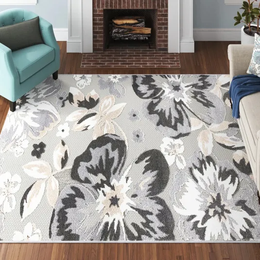A living room features a large area rug with a floral pattern in shades of gray, white, and beige, set on wooden flooring. An aqua armchair and light-colored sofa flank a brick fireplace.