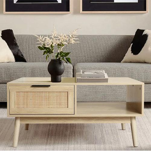 A modern wooden coffee table with open shelves and a rattan drawer is set in front of a gray sofa. The table holds a dark vase with white flowers and a neat stack of books on top.