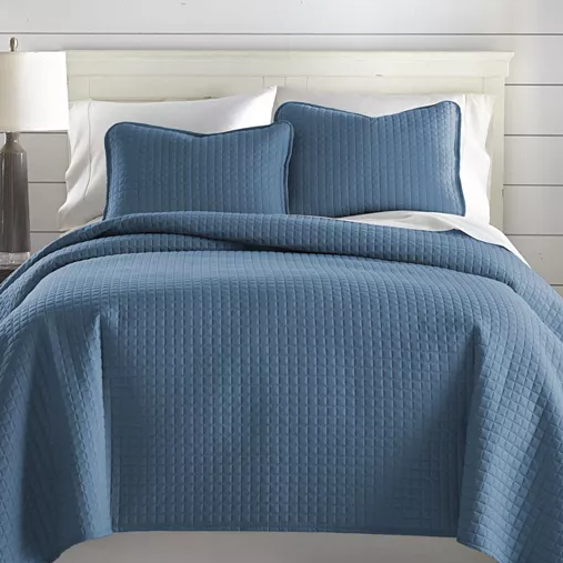 A blue 3-piece oversized queen quilt set featuring a textured, grid-like pattern, includes a quilt and two matching pillow shams. The quilt covers a white bed and is displayed in a well-lit room with a white headboard and a lamp on a side table.