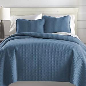 A blue 3-piece oversized queen quilt set featuring a textured, grid-like pattern, includes a quilt and two matching pillow shams. The quilt covers a white bed and is displayed in a well-lit room with a white headboard and a lamp on a side table.