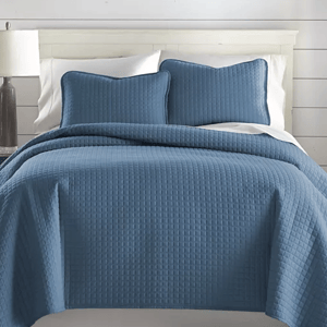 A blue 3-piece oversized queen quilt set featuring a textured, grid-like pattern, includes a quilt and two matching pillow shams. The quilt covers a white bed and is displayed in a well-lit room with a white headboard and a lamp on a side table.