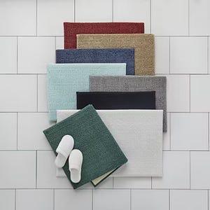 A collection of bath mats in various colors displayed on a tiled floor with a pair of white slippers on one mat.