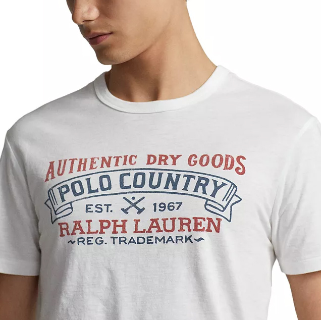 A white Polo Ralph Lauren men's t-shirt features a graphic design with the text \“Authentic Dry Goods Polo Country Est. 1967 Ralph Lauren Reg. Trademark\“ in red and blue.