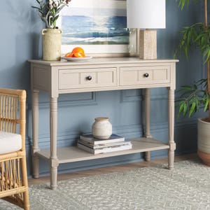 A beige console table features turned legs, two drawers, and a lower shelf, adorned with a lamp, vase, and decorative items.
