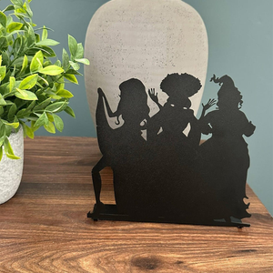 A silhouette decoration depicting three witch-like figures is shown, standing on a wooden surface next to a green potted plant and a large vase.