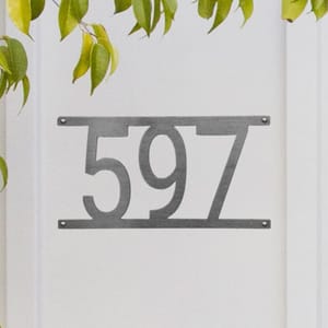 Brushed metal house number plaque displaying the numbers \“597\“ mounted on a white surface, with four visible screws and some leaves partially visible on the top left.