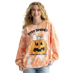 A woman wears an orange tie-dye sweatshirt featuring Snoopy sitting on a jack-o'-lantern with the text \“Stay Spooky\“.
