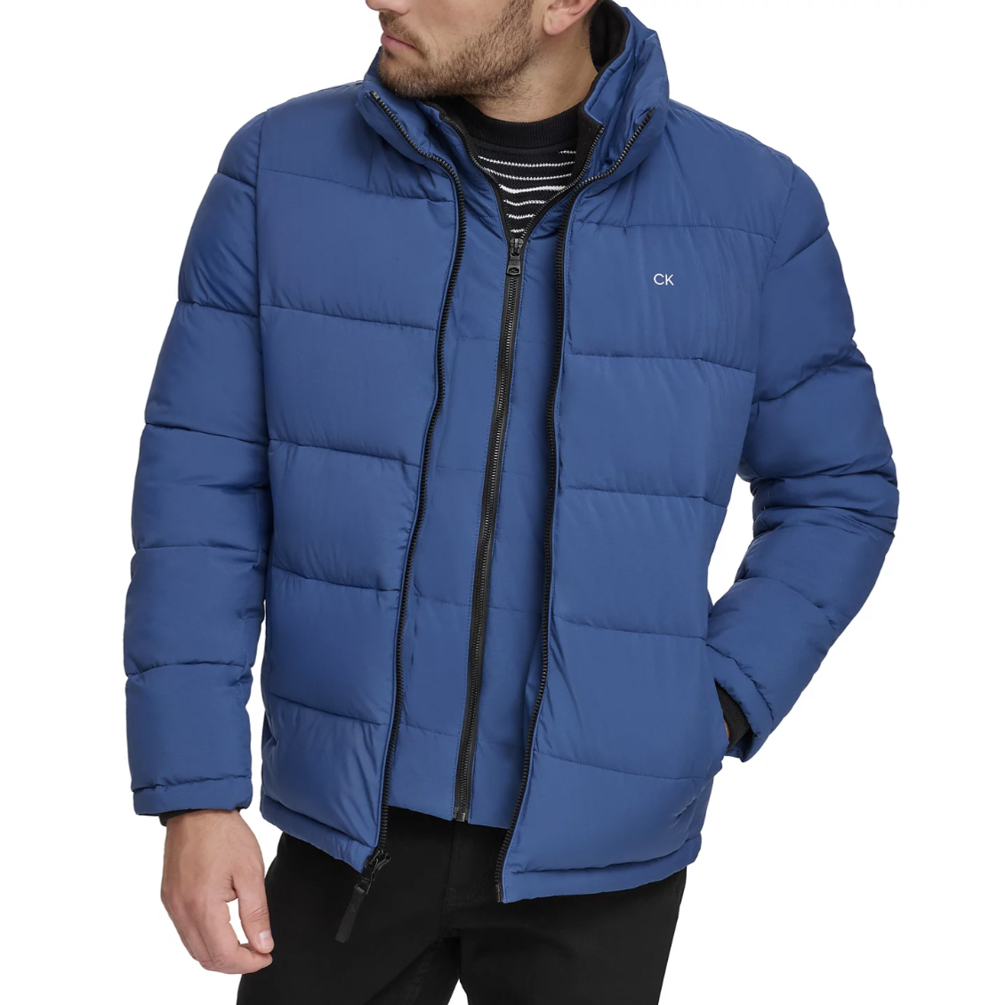 Blue puffer jacket with a zipper closure and CK logo, worn over a black and white striped shirt.
