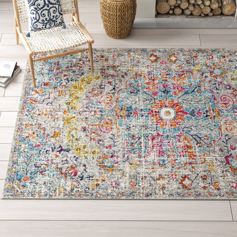 A colorful area rug with intricate patterns is placed on light wood flooring next to a woven chair with a white and blue patterned cushion, a small basket, and a stack of books.