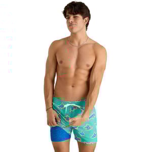 Chubbies Apex Swimmer Swim Trunks feature a green base with a colorful shark pattern and an asymmetric blue wave design on one leg. They have an adjustable waistband with a white drawstring.