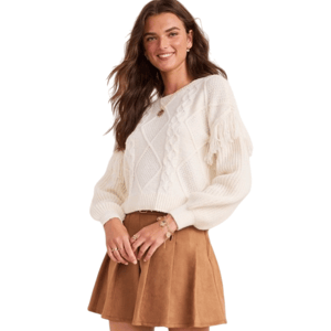 A woman models a white knit sweater with fringe details, paired with a brown suede mini skirt and tall brown leather boots. She accessorizes with a gold bracelet and necklace.