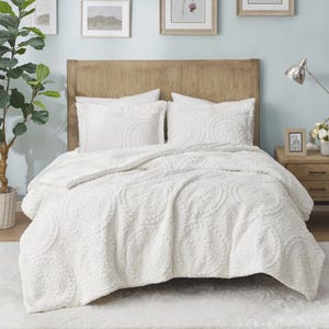 A cozy bedroom setup featuring a white textured quilt with circular patterns, matching shams, wooden headboard, wooden nightstands, and wall art. A floor lamp and indoor plant complement the decor.