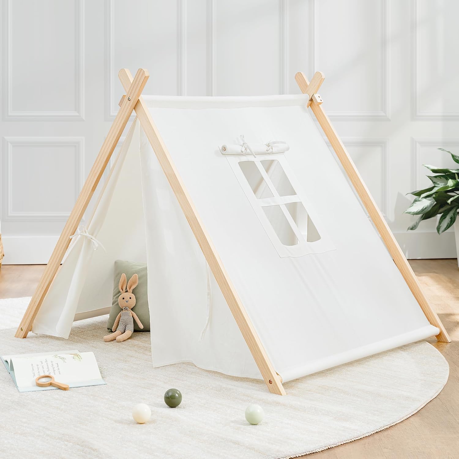 A children's play tent with a white fabric cover, natural wood frame, and a small window at the front. Inside, there's a plush rabbit toy and a cushion, with a book and three balls on the soft, round rug outside.
