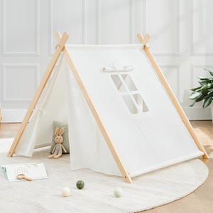 A children's play tent with a white fabric cover, natural wood frame, and a small window at the front. Inside, there's a plush rabbit toy and a cushion, with a book and three balls on the soft, round rug outside.