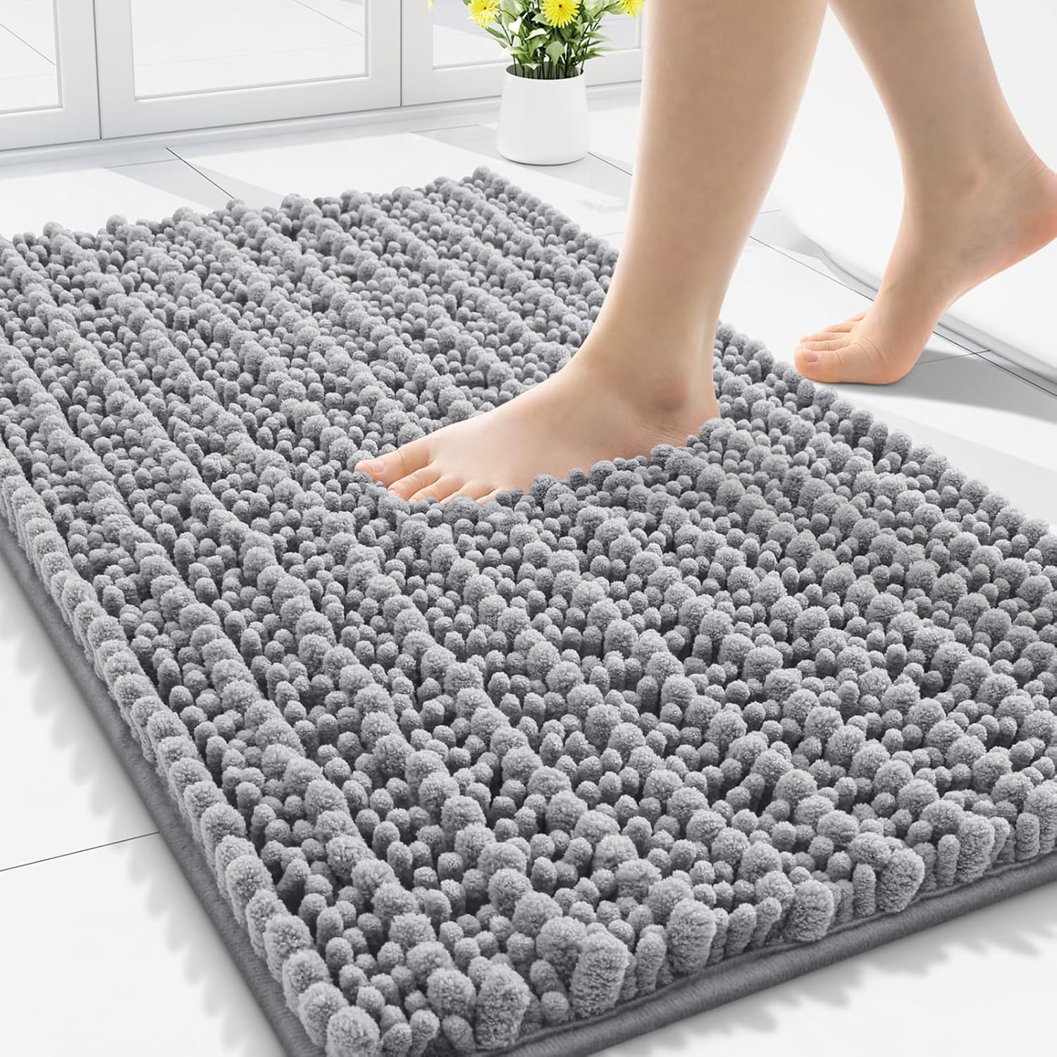 A gray, textured bath mat with a soft, shaggy surface, shown being stepped on with bare feet. A white tile floor and a yellow flowering plant are in the background.