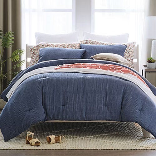 A neatly arranged bed features a blue comforter, multiple pillows with varying designs, and a patterned throw blanket. The setup is complemented by a green plant and wooden bedside table.