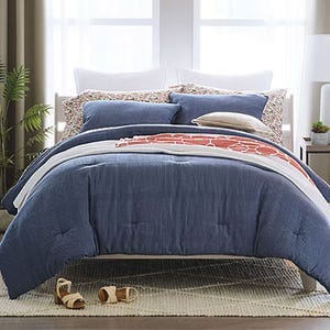 A neatly arranged bed features a blue comforter, multiple pillows with varying designs, and a patterned throw blanket. The setup is complemented by a green plant and wooden bedside table.