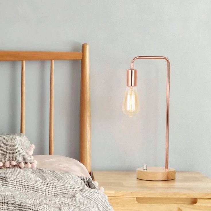 A modern bedside table features a sleek copper lamp with an exposed bulb, a wooden base, and is complemented by a wooden bedframe and a textured blanket with pom-pom fringes.