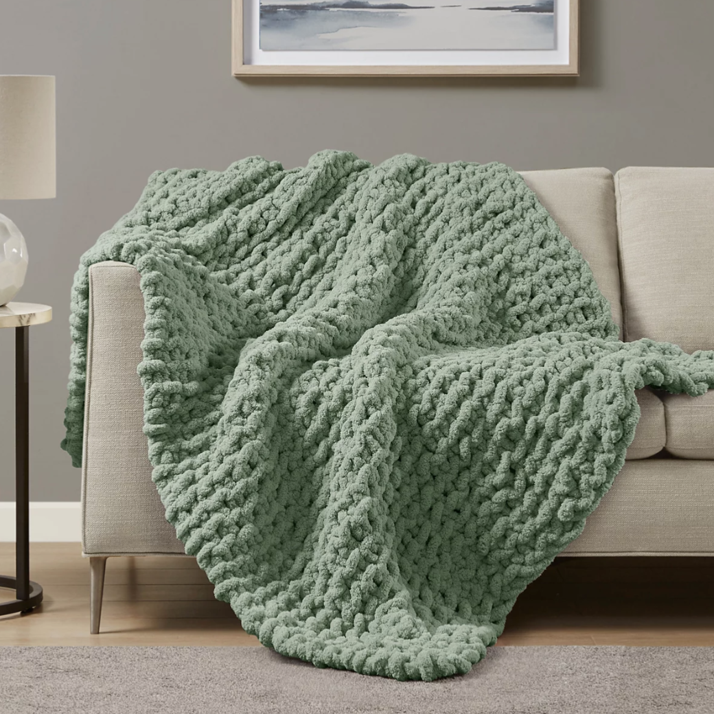 A mint green chunky knit throw draped over a light beige couch, with a minimalist landscape artwork above and a lamp on the side table.