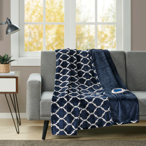 A navy and white patterned heated blanket with a remote drapes over a gray sofa, next to a small side table with a potted plant and lamp, against a window backdrop with an autumn view.