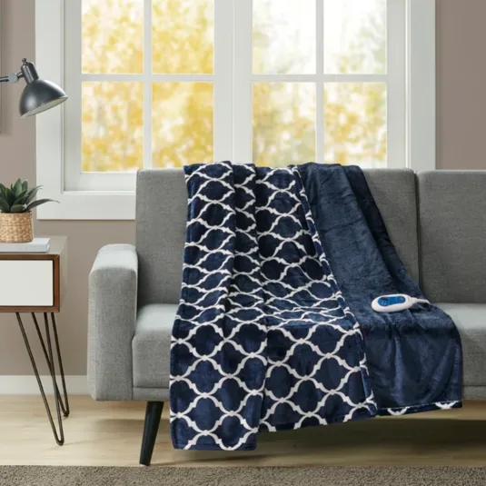 A navy and white patterned heated blanket with a remote drapes over a gray sofa, next to a small side table with a potted plant and lamp, against a window backdrop with an autumn view.