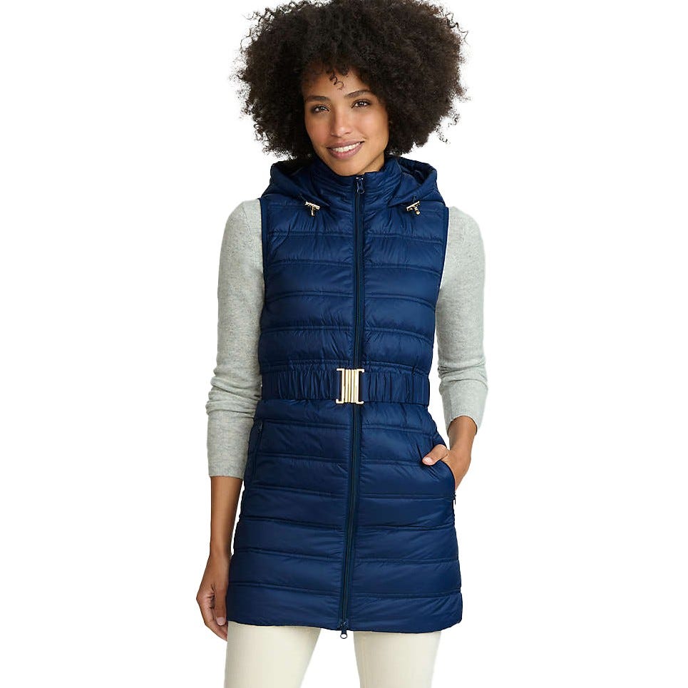 A woman is wearing a blue quilted hooded vest with a gold buckle belt, over a light gray long-sleeve top and white pants.