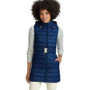 A woman is wearing a blue quilted hooded vest with a gold buckle belt, over a light gray long-sleeve top and white pants.