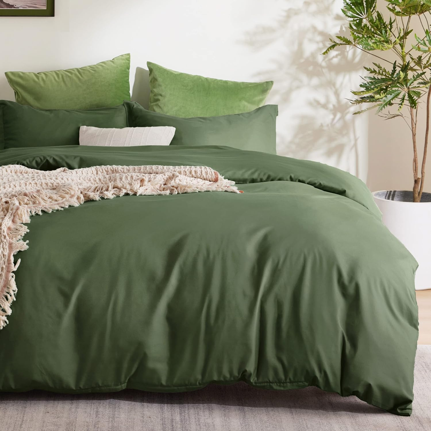Green bedding set with duvet cover and pillows, accented with a white decorative pillow and a textured beige throw blanket, accompanied by a potted plant in a room setting.