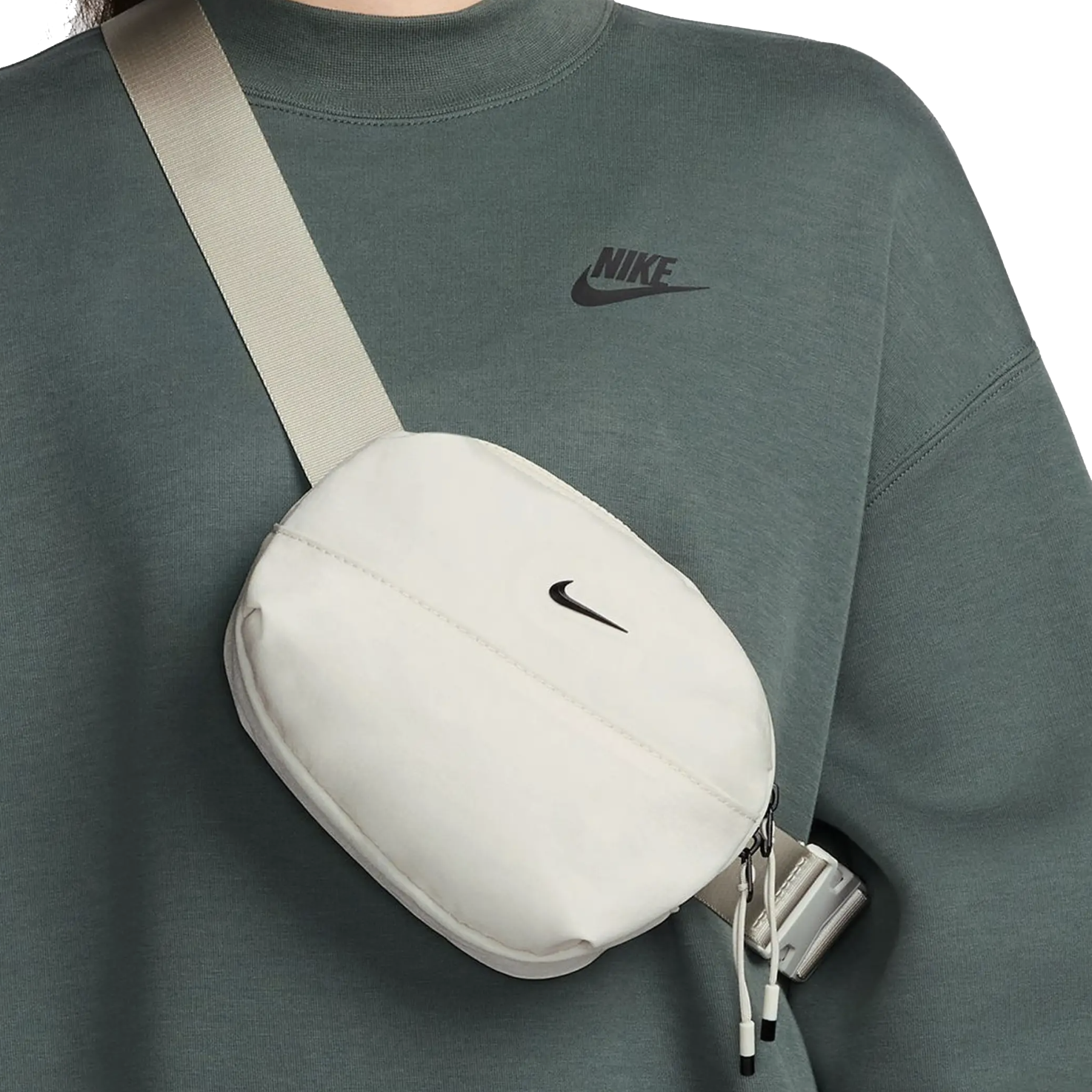 A small, white Nike belt bag with the logo on the front, featuring a zippered closure and an adjustable strap worn across the chest. It's paired with a dark green Nike sweatshirt displaying the brand's logo.
