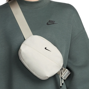 A small, white Nike belt bag with the logo on the front, featuring a zippered closure and an adjustable strap worn across the chest. It's paired with a dark green Nike sweatshirt displaying the brand's logo.