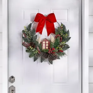 A festive wreath featuring a house motif, decorated with a red bow, pinecones, greenery, and red accents, hangs on a white door.