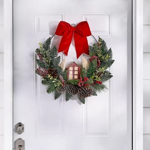 A festive wreath featuring a house motif, decorated with a red bow, pinecones, greenery, and red accents, hangs on a white door.