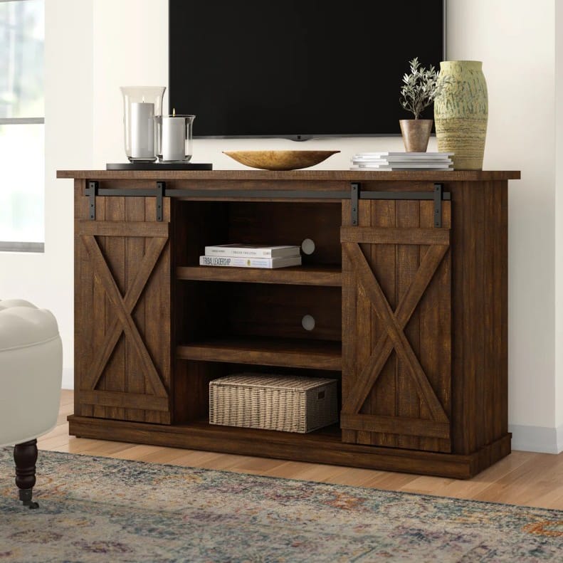 Rustic wooden TV stand with sliding barn doors, shelves for storage, and a woven basket. Decor includes candles, books, a wooden bowl, and a vase with a plant.