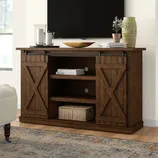 Rustic wooden TV stand with sliding barn doors, shelves for storage, and a woven basket. Decor includes candles, books, a wooden bowl, and a vase with a plant.