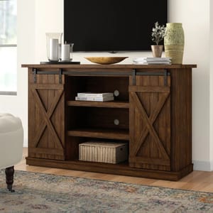 Rustic wooden TV stand with sliding barn doors, shelves for storage, and a woven basket. Decor includes candles, books, a wooden bowl, and a vase with a plant.