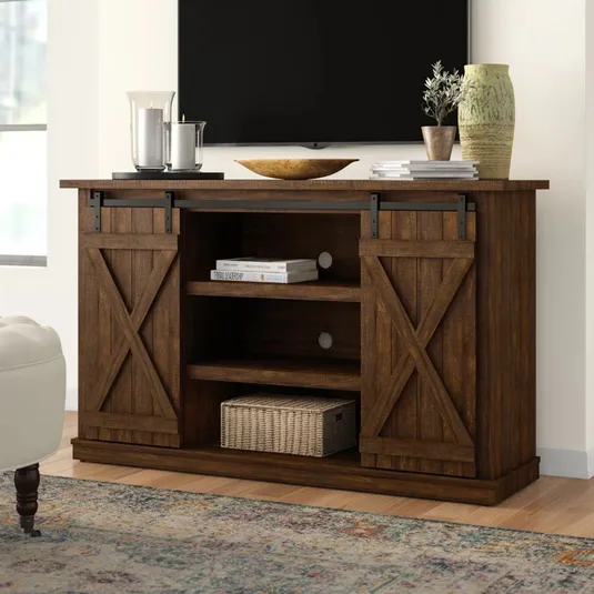 Rustic wooden TV stand with sliding barn doors, shelves for storage, and a woven basket. Decor includes candles, books, a wooden bowl, and a vase with a plant.