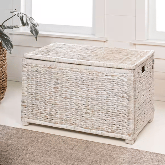 A rectangular wicker storage chest with a light whitewashed finish, featuring woven texture and side handles, is placed on a carpeted floor near a window and greenery.