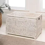 A rectangular wicker storage chest with a light whitewashed finish, featuring woven texture and side handles, is placed on a carpeted floor near a window and greenery.