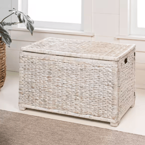 A rectangular wicker storage chest with a light whitewashed finish, featuring woven texture and side handles, is placed on a carpeted floor near a window and greenery.