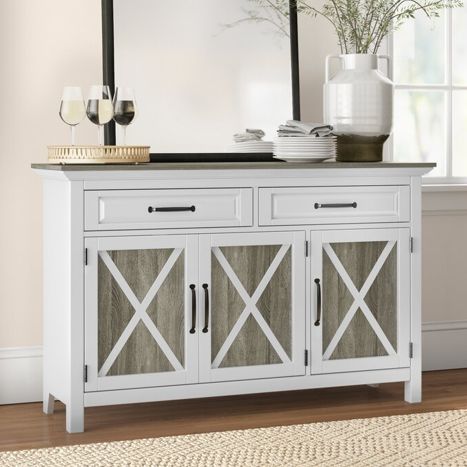 A white 52\“ farmhouse sideboard with two drawers and three cabinet doors featuring an X-pattern design. It has black handles and is topped with glassware and kitchen items, complementing a rustic decor style.