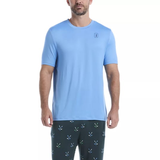 A man is wearing a light blue short-sleeve T-shirt with a small logo on the chest and dark pants featuring green and blue crossed designs.