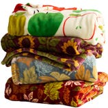 A stack of four colorful, patterned fleece blankets featuring designs with apples, sunflowers, autumn leaves, and more leaves on a red background.