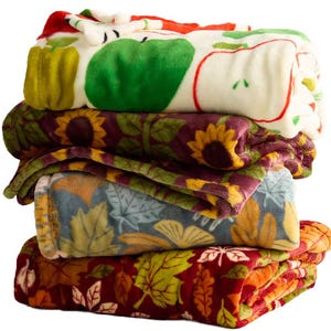 A stack of four colorful, patterned fleece blankets featuring designs with apples, sunflowers, autumn leaves, and more leaves on a red background.