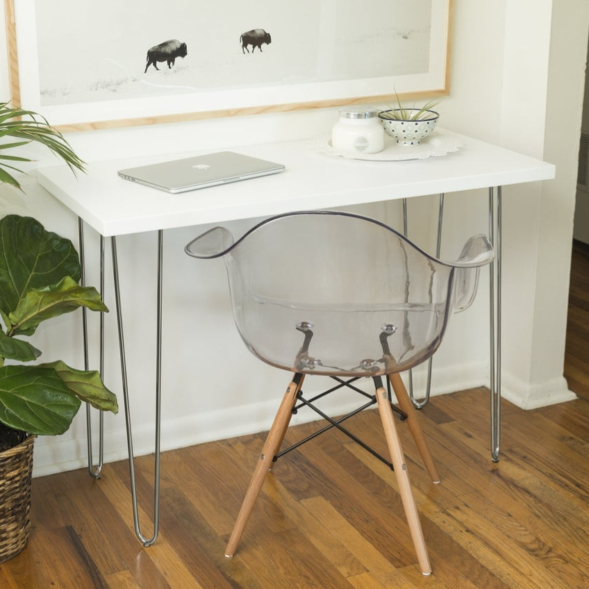 A minimalist workspace featuring a white desk with hairpin legs, a transparent plastic chair with wooden legs, a laptop, a potted plant, and a black-and-white framed photo of bison, set on a wooden floor.