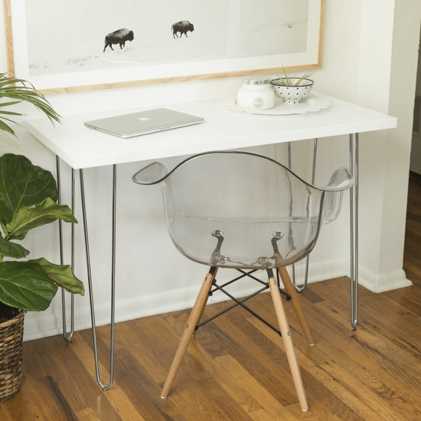 A minimalist workspace featuring a white desk with hairpin legs, a transparent plastic chair with wooden legs, a laptop, a potted plant, and a black-and-white framed photo of bison, set on a wooden floor.