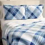 A bed is neatly made with blue and white plaid patterned pillows and matching blanket, complemented by a white sheet underneath.