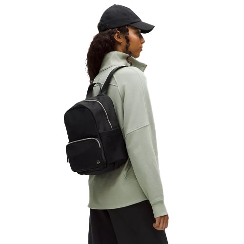 A person wearing a light green jacket, black cap, and black backpack with silver zippers.