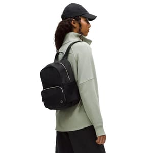 A person wearing a light green jacket, black cap, and black backpack with silver zippers.